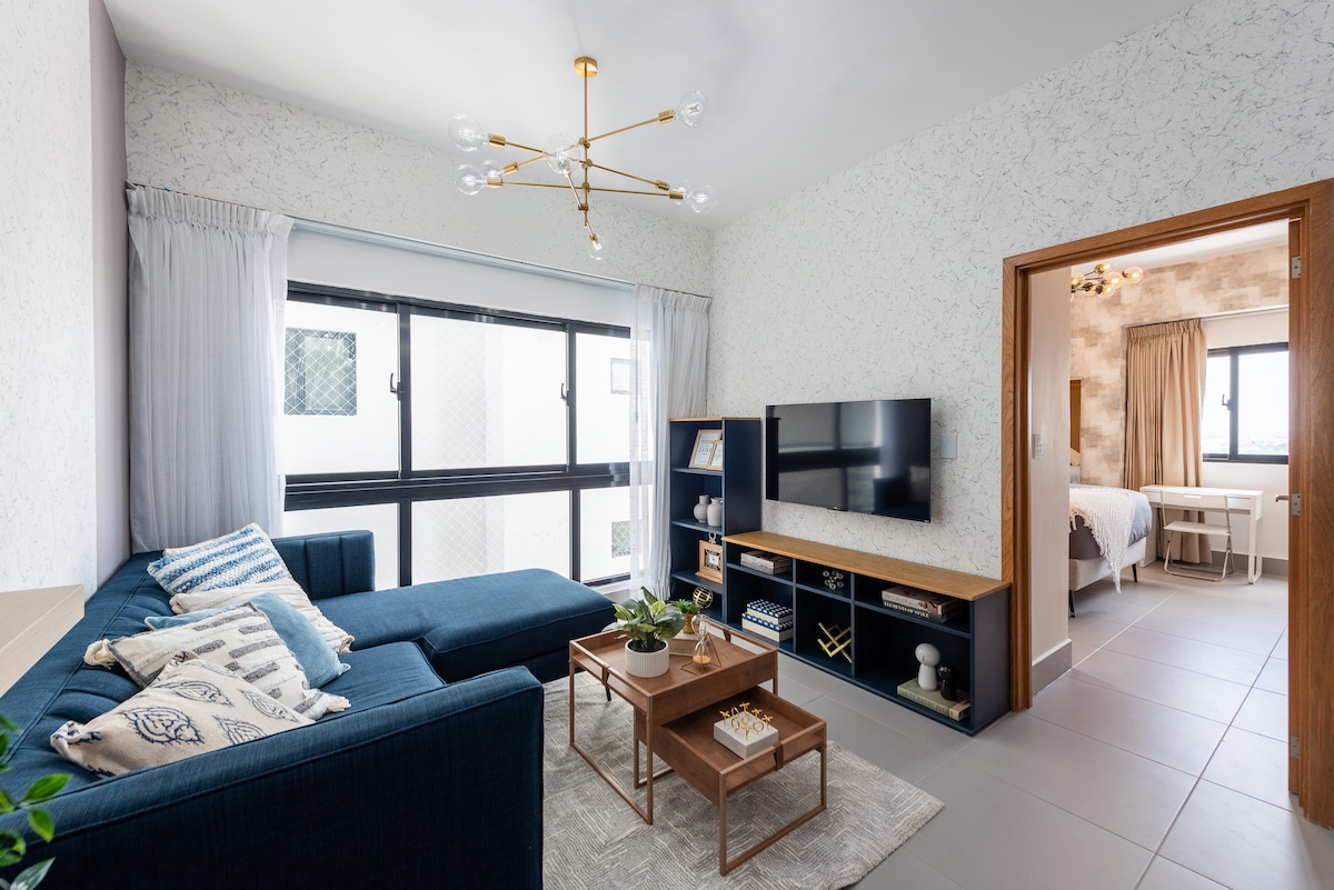 A modern living room is featured, characterized by a sectional sofa and a wooden coffee table. A flat-screen television is mounted on the wall. Natural light enters through large windows adorned with sheer curtains, allowing glimpses of the adjoining bedroom.