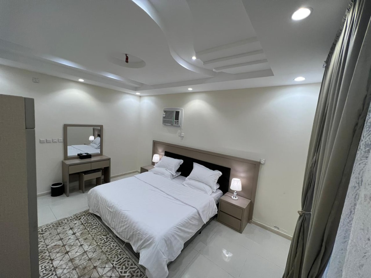 A spacious sleeping area is presented with a large bed featuring soft white linens. Two bedside tables with lamps are positioned on either side of the bed, and a dressing mirror is situated against the wall. Soft lighting enhances the calm ambiance.