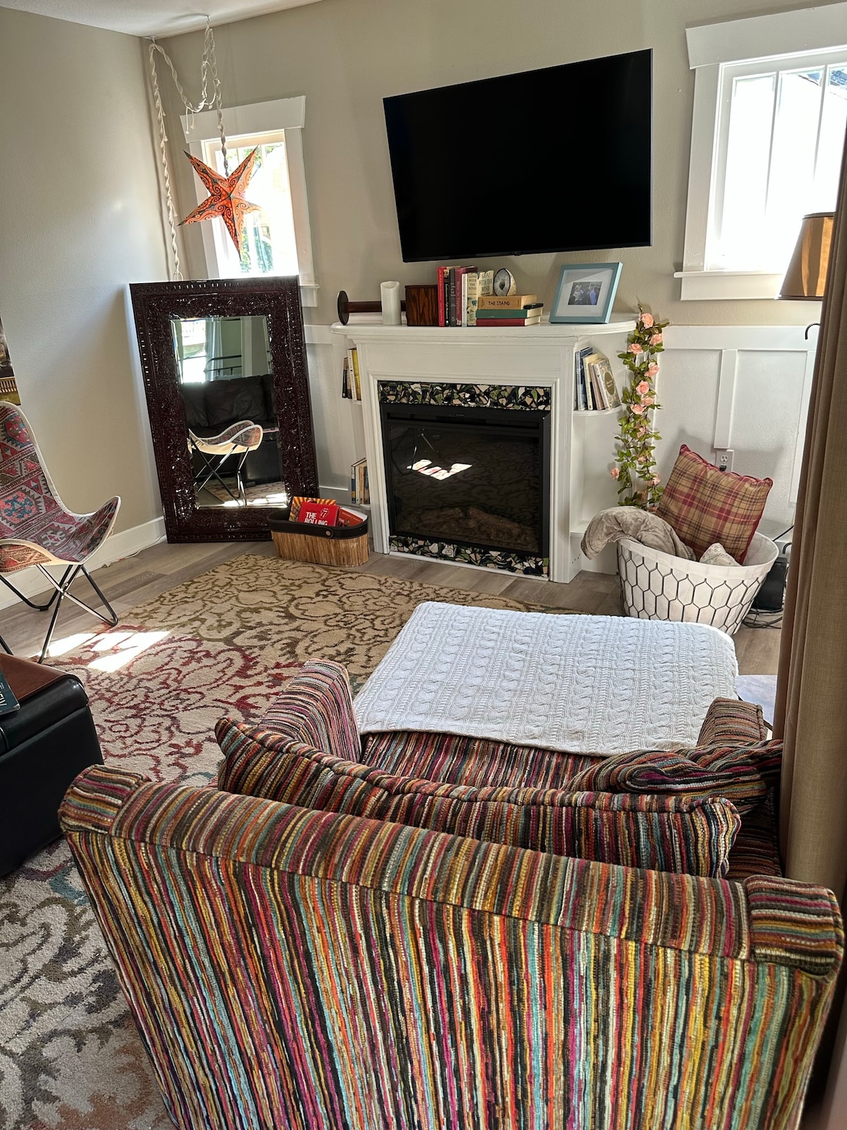 A cozy living area features a striped armchair facing a decorative fireplace with a mirror above it. A large TV is mounted on the wall, and natural light filters in through the windows. A woven basket and patterned rug add texture to the space.
