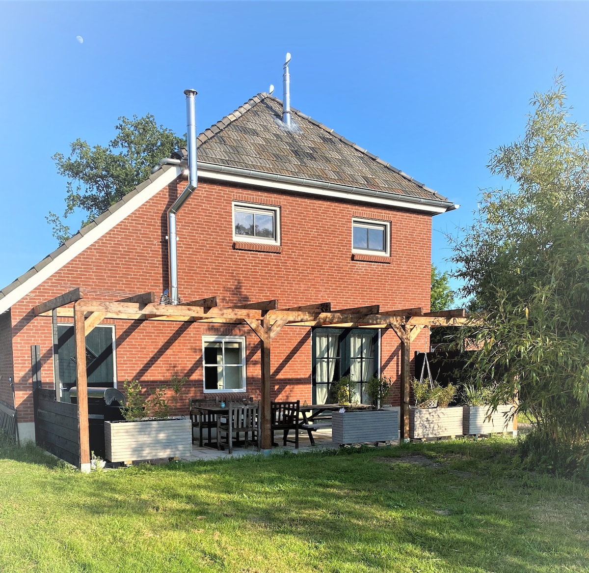 The exterior of the spacious red-brick farmhouse is highlighted by a wooden pergola, providing shade for the outdoor seating area. Lush green grass surrounds the property, and a few windows reflect natural light. A clear blue sky above creates an inviting atmosphere.