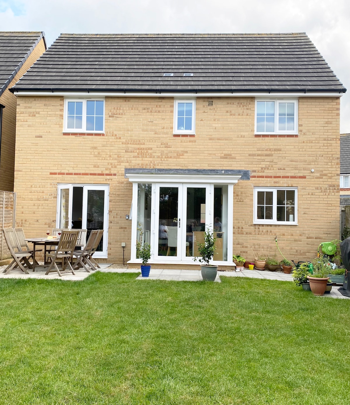 A two-story modern home is set within a well-kept garden. Large glass doors provide access to the outdoor patio area, furnished with multiple wooden chairs and a table. Lush green grass surrounds the home, complemented by potted plants and a tidy walk path.