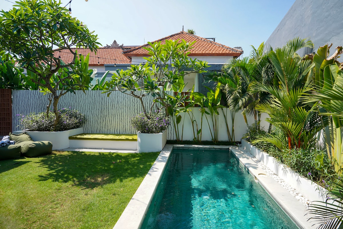 A private pool is framed by lush tropical plants and landscaped gardens, enhancing the serene outdoor environment. The clear water reflects the blue sky, and a small seating area with cushions is visible on the manicured lawn, inviting relaxation.