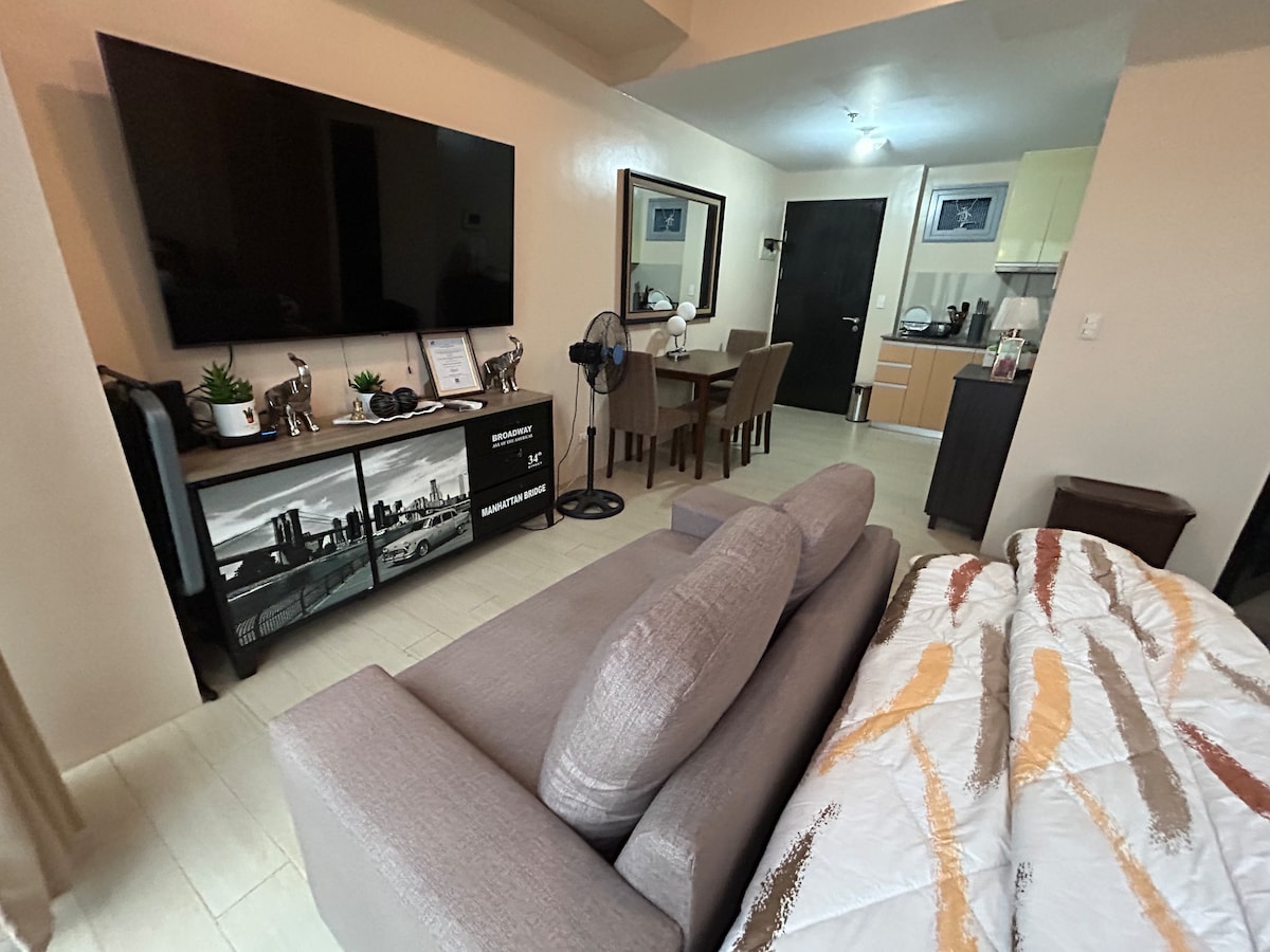 A well-organized space features a cozy sofa facing a large flat-screen TV and a stylish media console. A dining table is visible, accompanied by four chairs. The kitchen area is accessible, providing a functional layout. Two mirrors enhance the sense of openness.