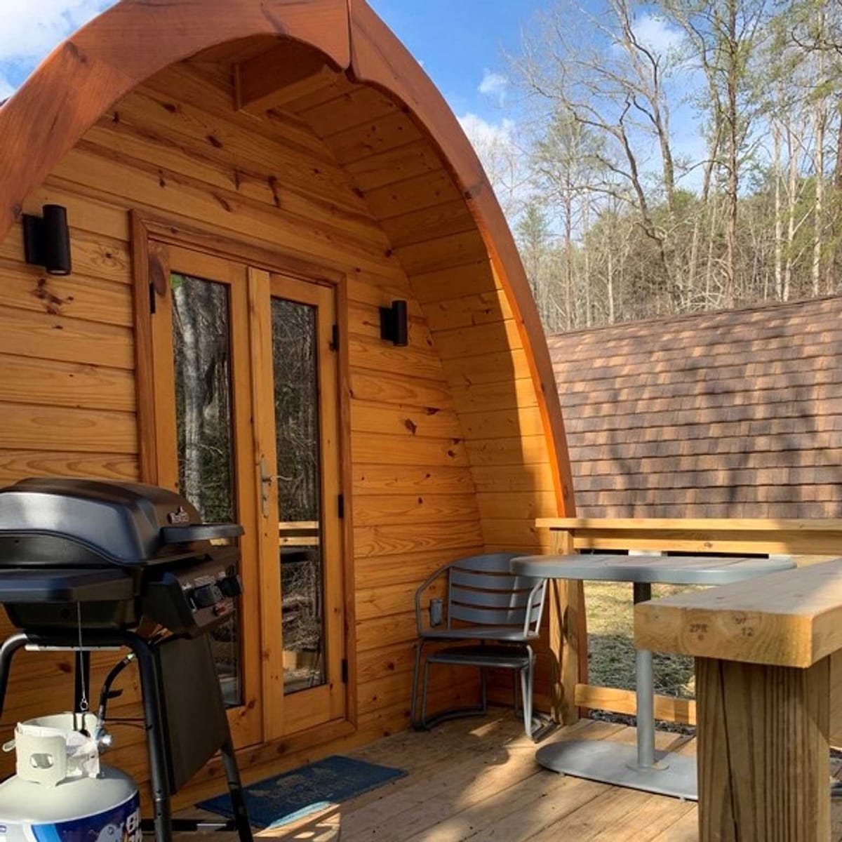 An outdoor deck features wooden walls with a curved roof, enhancing the cozy feel. A barbecue grill is positioned next to a table and a chair, inviting outdoor meals. Natural scenery can be seen in the background, contributing to a serene environment.