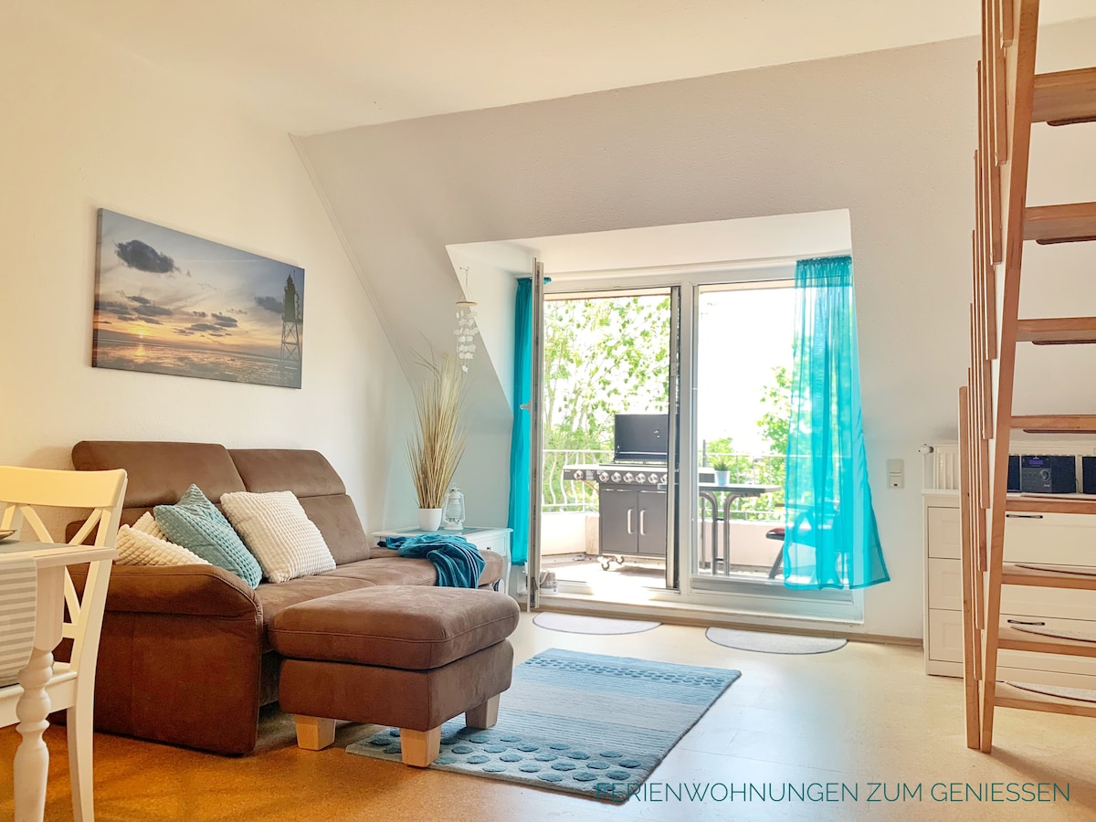 A bright living area features a comfortable brown sofa, accompanied by a light blue area rug. A dining table with white chairs sits nearby. Large glass doors open to a balcony with greenery, with a gas grill visible outside.