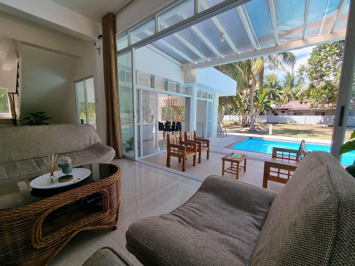 A bright living area is seen through large glass doors that open to the patio and pool. Comfortable seating is arranged around a small table. Natural light floods the space, highlighting the connection between indoor and outdoor areas surrounded by greenery.