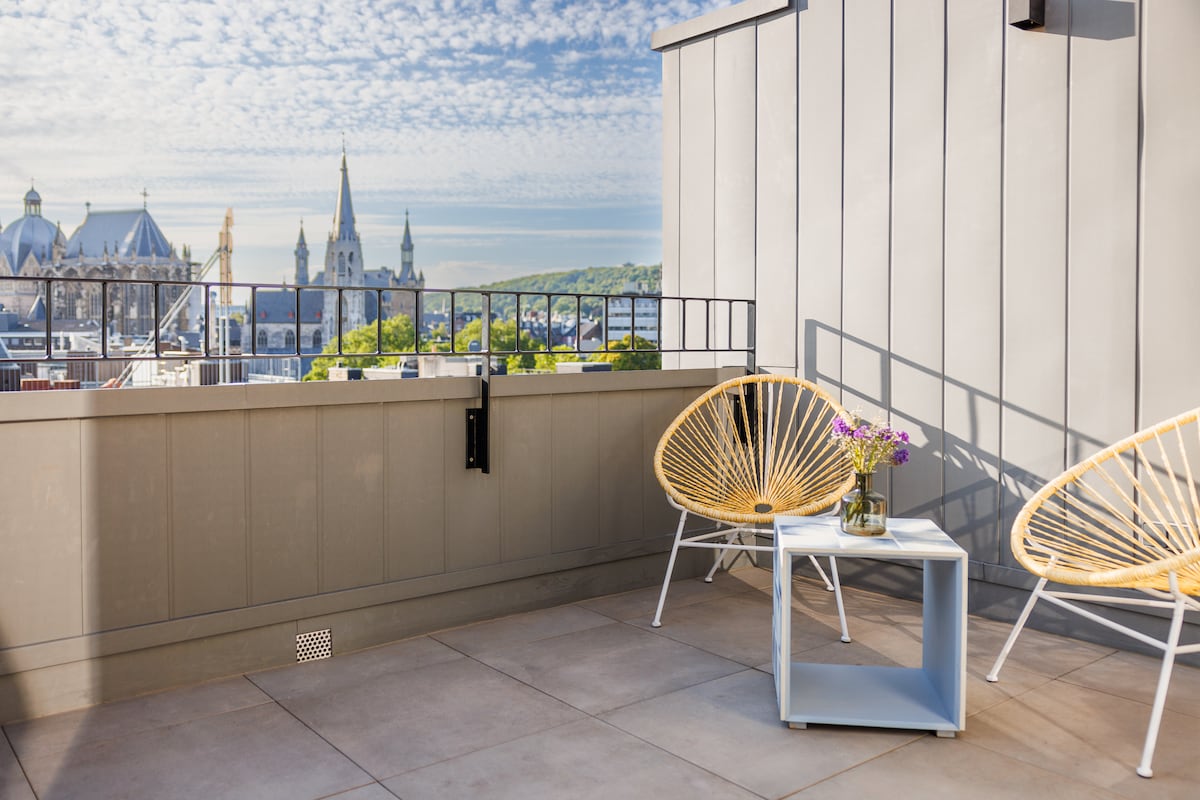 A sunlit terrace is displayed, featuring two round, woven chairs beside a small, light-colored table. A vase with fresh flowers rests on the table. The background offers a scenic view of the skyline, highlighting historic architecture and a clear blue sky.