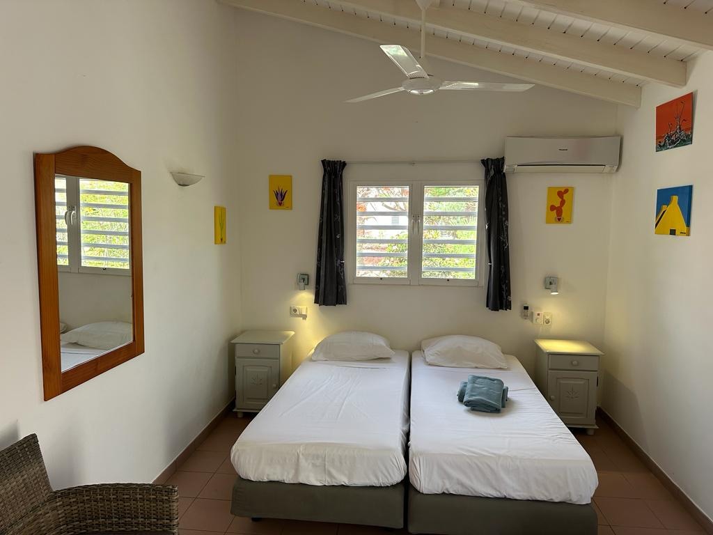 The studio features two single beds with fresh linen arranged neatly. A mirror and bedside tables are positioned beside the beds. Natural light filters through the partially open windows, and colorful artwork decorates the white walls, enhancing the room's bright atmosphere.