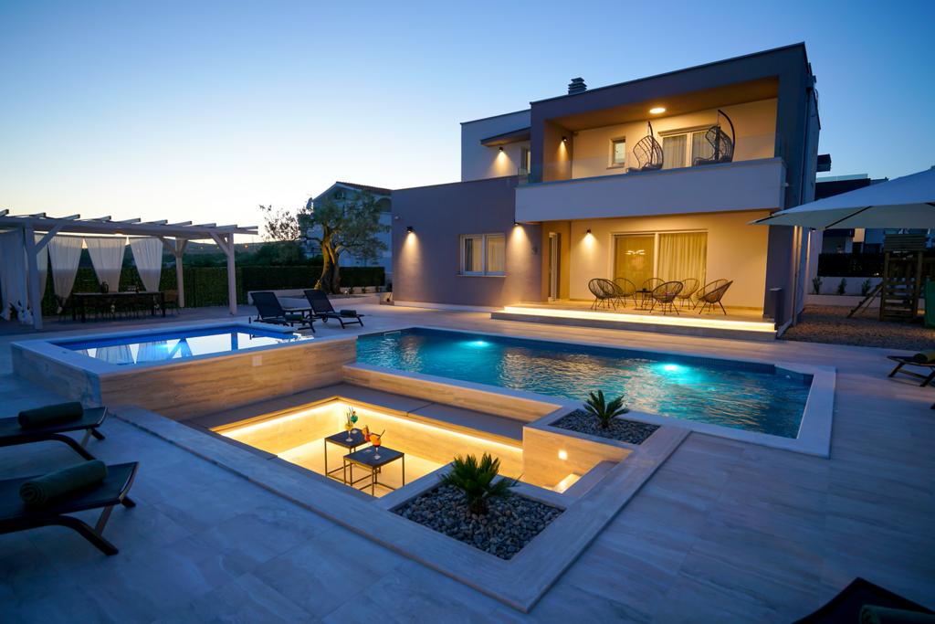 An outdoor area is presented featuring two swimming pools, surrounded by a spacious sun deck. Lounge chairs are positioned nearby. Soft lighting illuminates the pools, enhancing the modern architectural design of the villa in the background during twilight.