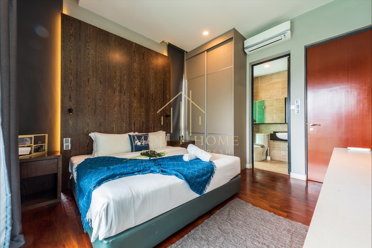 A spacious bedroom features a comfortable bed with a dark blue throw draped over it. Wooden accents adorn the wall behind, while natural light enters through a nearby window. A small bedside table is positioned next to the bed, offering a minimalist decor.