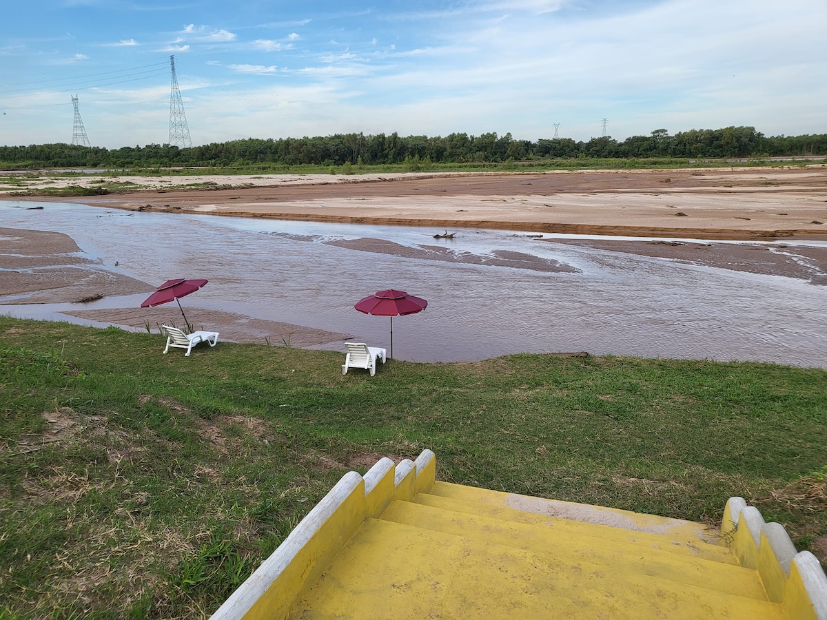 Two lounge chairs are positioned beside a gently flowing river, shaded by maroon umbrellas. The grassy area extends towards the water, which reflects the sky. In the background, a natural landscape is visible, bordered by low foliage and distant power lines.
