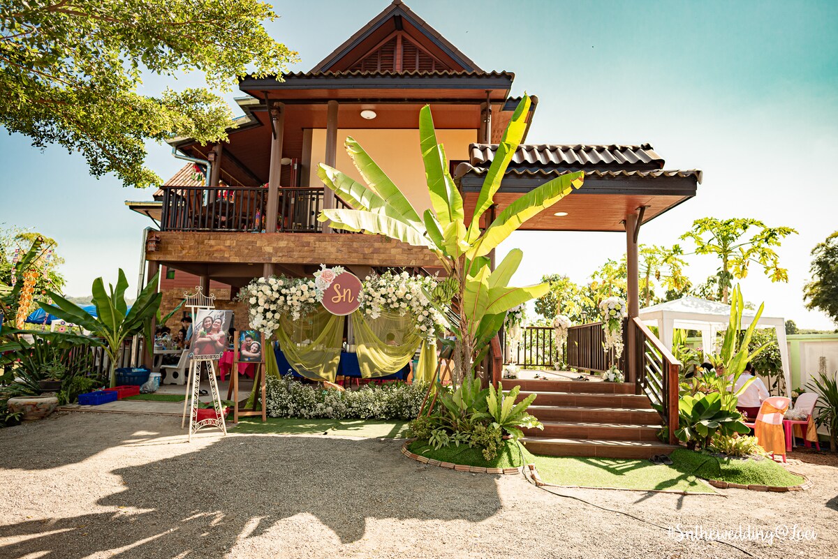 A charming two-story house is surrounded by lush greenery and tropical plants. The entrance features a set of wooden stairs leading to a covered porch with seating, framed by vibrant flowers and decorative elements, inviting guests to enjoy the outdoor space.