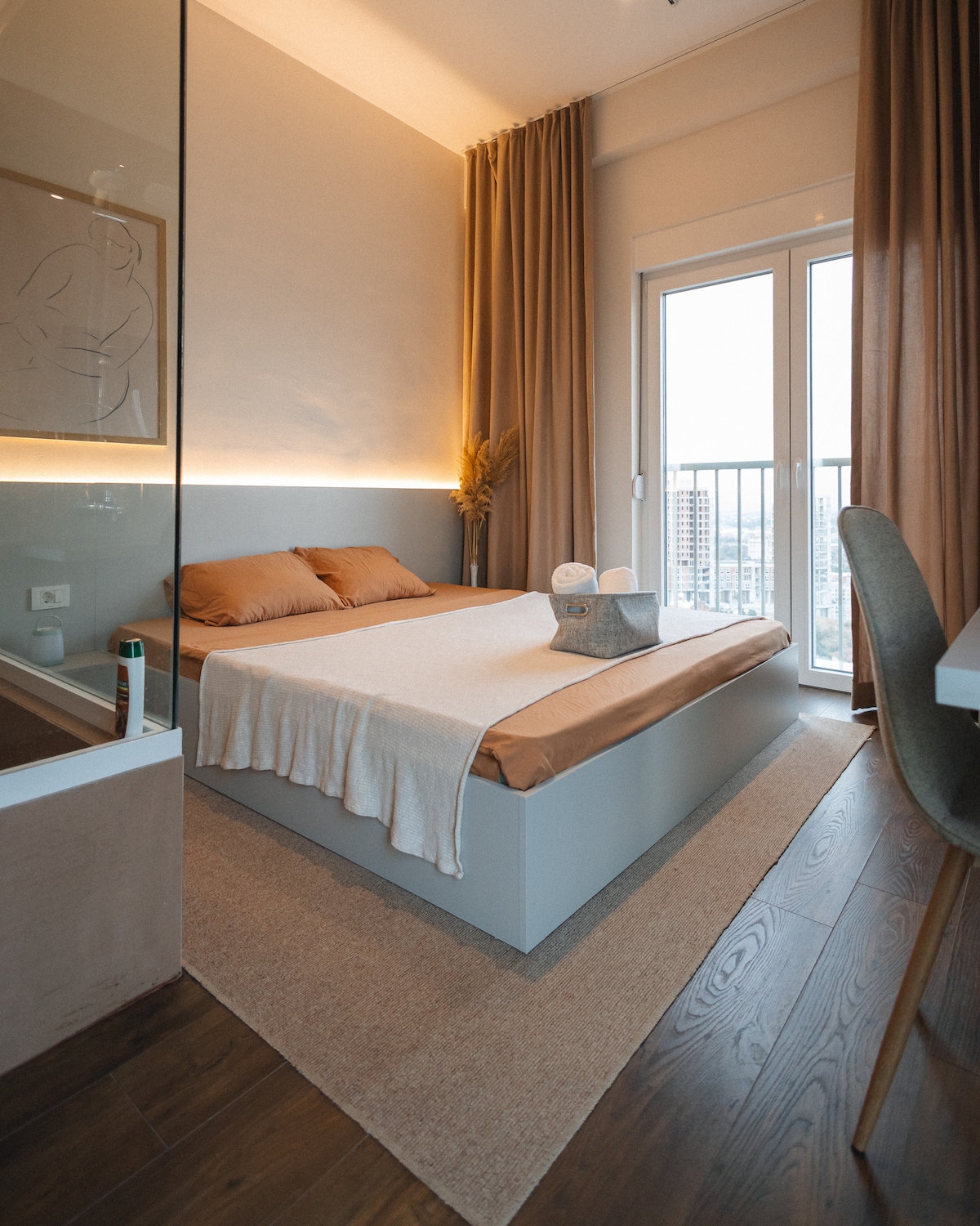 A cozy bedroom features a spacious bed dressed in neutral linens, complemented by warm-toned pillows. Large windows allow natural light to fill the space, while soft curtains provide privacy. A simple chair and desk offer additional functionality, creating a serene atmosphere for relaxation.