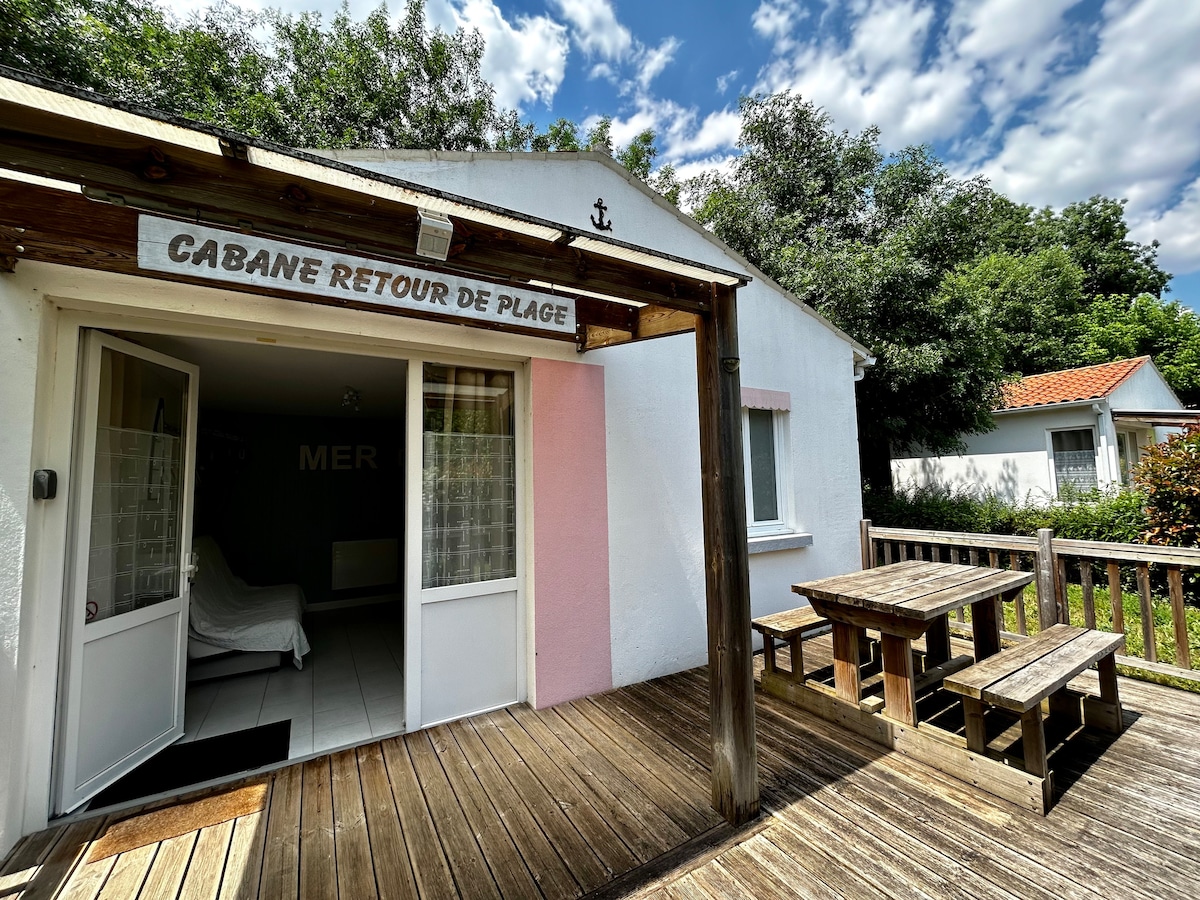 A wooden deck creates an inviting space in front of the cabin, featuring a picnic table for outdoor dining. The cabin's entrance is adorned with a sign reading 'Cabane Retour de Plage', while lush greenery surrounds the area, contributing to a serene environment.