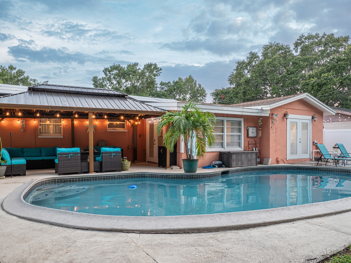 An inviting outdoor living space is depicted, featuring a refreshing pool surrounded by a concrete patio. Lush palm trees provide a tropical feel, while comfortable seating is arranged under a covered area. Soft lighting adorns the setting, enhancing the relaxed atmosphere.