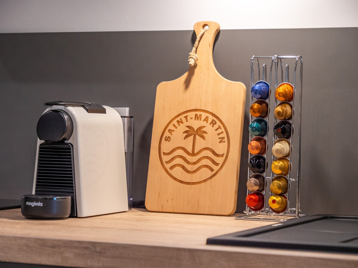 A modern kitchen countertop features an elegant coffee machine alongside a decorative wooden cutting board engraved with 'SAINT-MARTIN.' A chrome stand displays a selection of colorful coffee capsules, providing convenience for coffee enthusiasts.