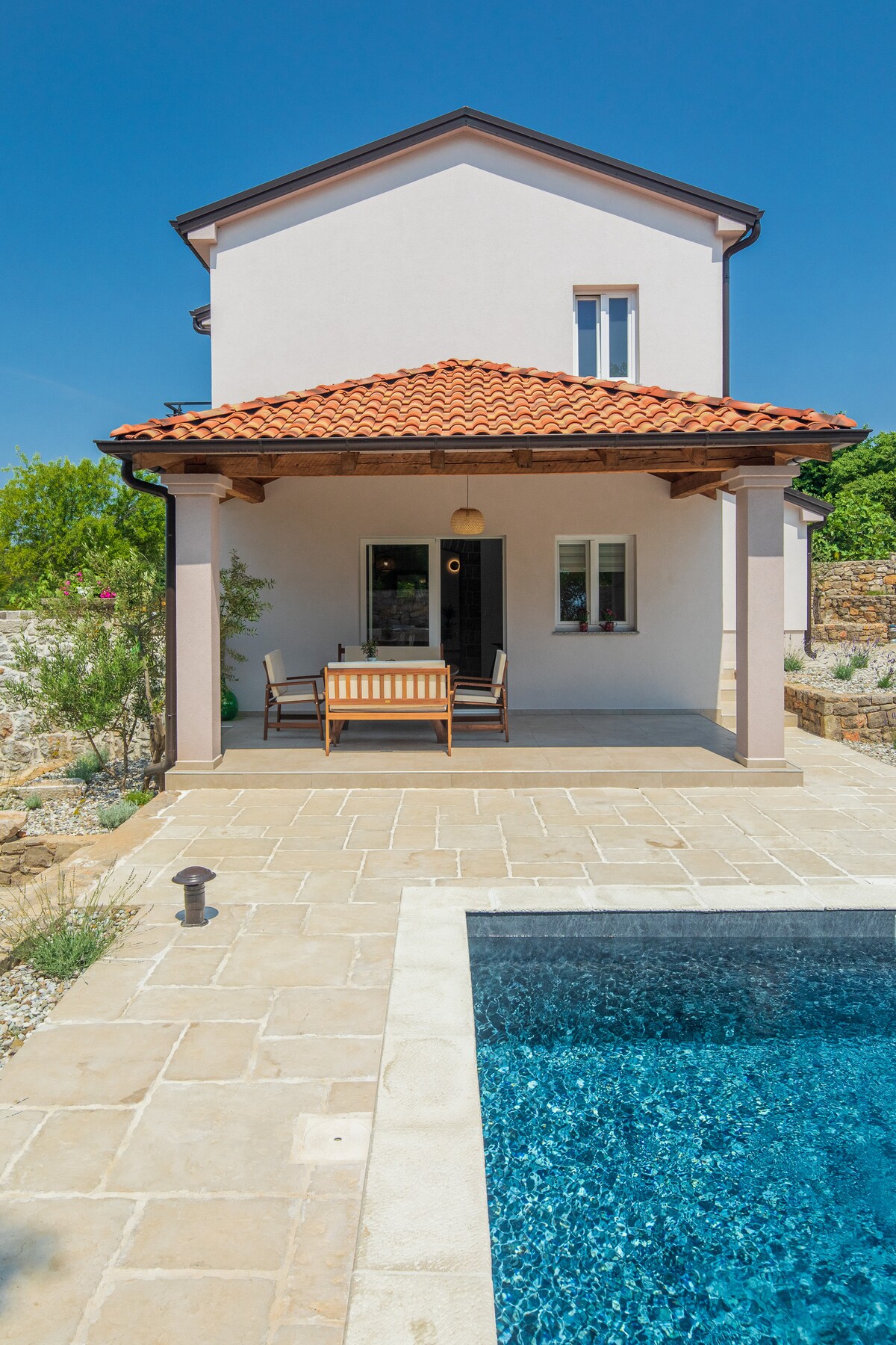 The exterior of the villa is shown, featuring a two-story structure with a red-tiled roof and stone patio. A small seating area with wooden chairs is visible under the covered porch, adjacent to a swimming pool that reflects clear blue water.