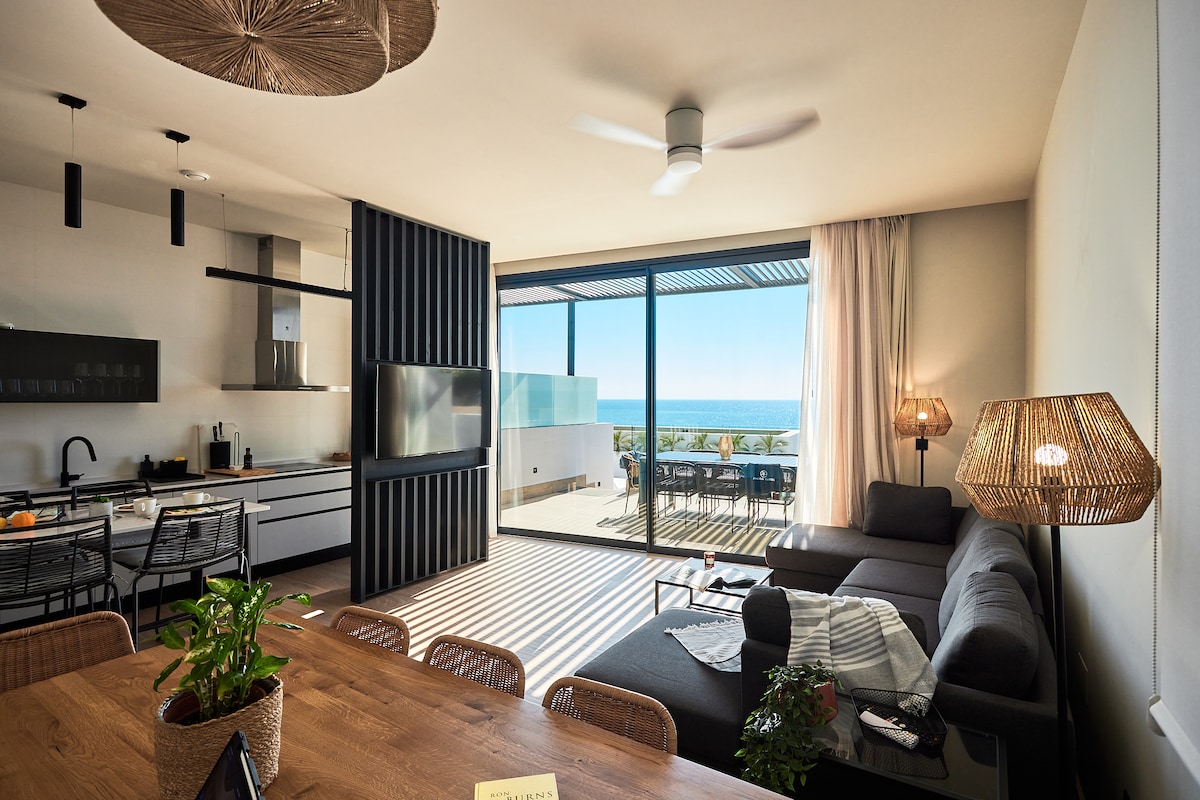 A modern living area features a comfortable sectional sofa and dining table with woven chairs. Large glass doors provide a view of the ocean and access to an outdoor terrace. Soft natural light filters through sheer curtains, enhancing the open space.