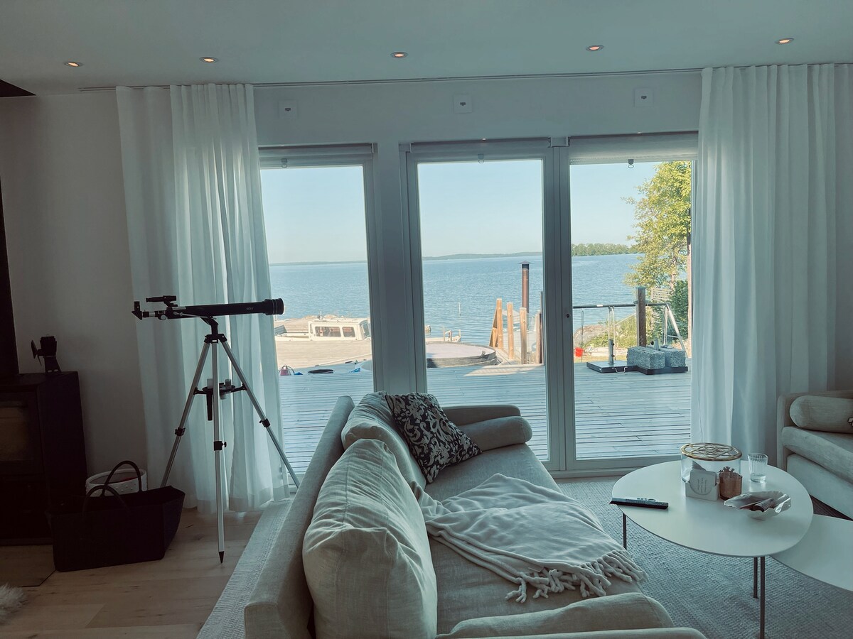 A comfortable living space features soft, light-colored sofas arranged around a low coffee table. Large glass doors provide a clear view of the serene waterfront, with a telescope positioned nearby for observation. Natural light filters through sheer curtains, enhancing the inviting atmosphere.