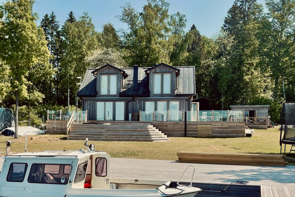 A modern two-story house is set among trees, featuring large glass doors that open to a spacious deck. The home’s exterior combines wood and metal elements, while the surrounding lawn provides plenty of green space. A small boat is anchored nearby, ready for exploration.