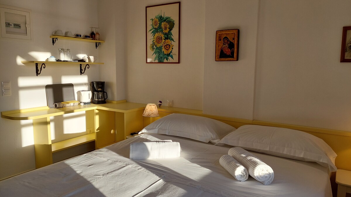 A bright room is presented, featuring a spacious bed with white linens neatly arranged. Two rolled towels are placed on the bed. A small desk area with coffee-making equipment is visible along the wall. Soft light filters through a nearby window, creating a warm atmosphere.