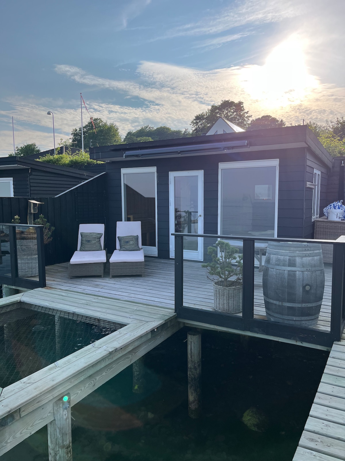 The exterior of the beach cabin features a spacious deck with two lounge chairs positioned for relaxation. A large glass door leads into the cabin, while a potted plant and a barrel add natural elements to the space. Sunlight creates a warm glow over the scene.