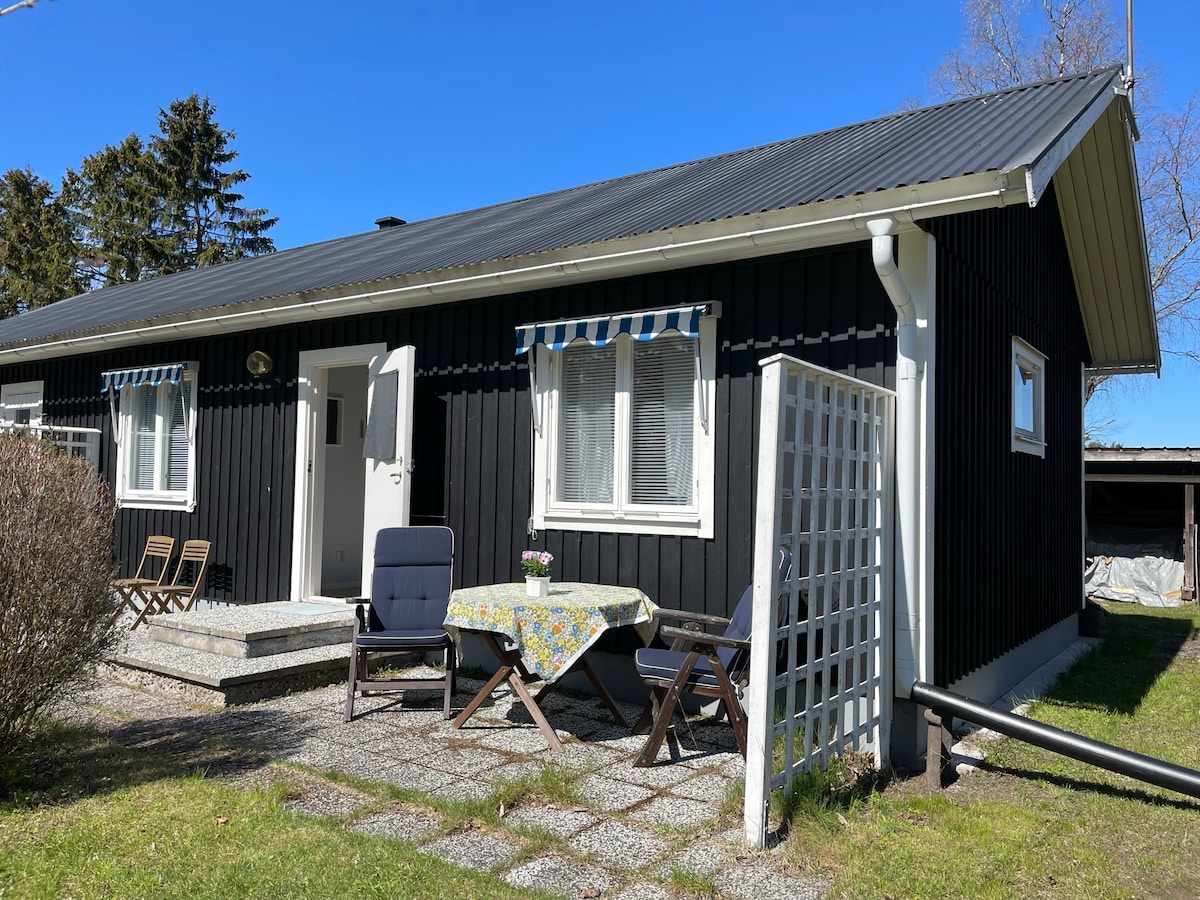 A black wooden cottage stands under a clear blue sky, complemented by a covered entrance. A small outdoor table adorned with a floral tablecloth is set between two chairs. Green grass surrounds the stone pathway leading to the front door, adding a touch of nature to the space.