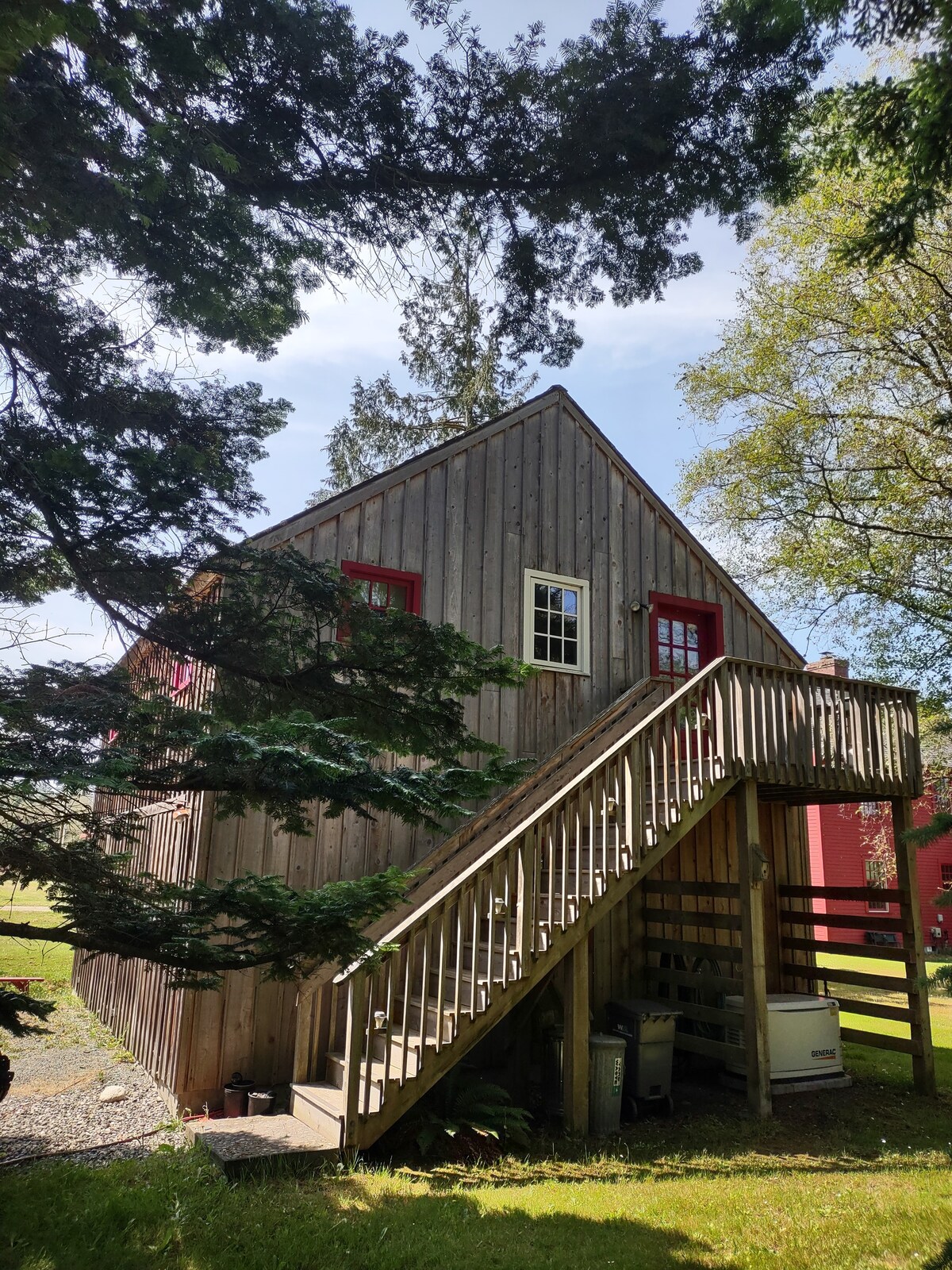 A wooden structure is positioned among trees, featuring a sloped roof and red-trimmed windows. Access to the studio apartment is provided by a wooden staircase leading to a deck. A grassy area surrounds the base of the building, contributing to a serene environment.