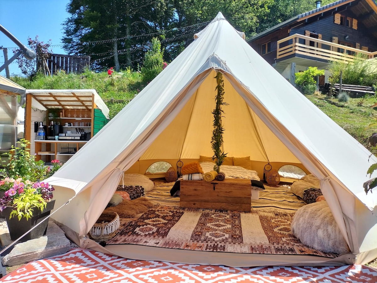A spacious glamping tent is set up with a central wooden table surrounded by pillows on the floor. Colorful textiles cover the ground, and plants enhance the serene atmosphere. The tent's entrance opens to a scenic outdoor space, framed by lush greenery.