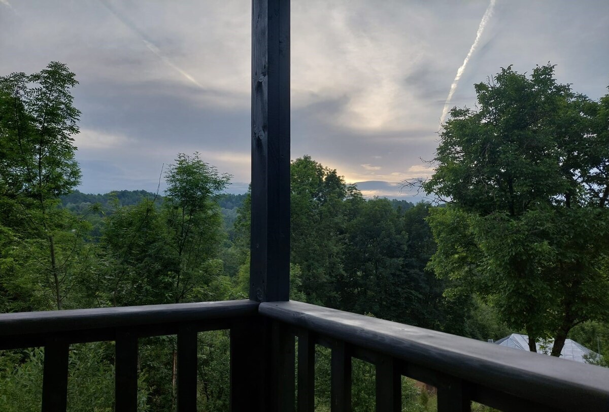 A view from the terrace showcases the surrounding lush greenery and distant hills under a soft sky. The horizon is marked by gentle clouds, creating a serene and expansive natural setting, framed by wooden railings.