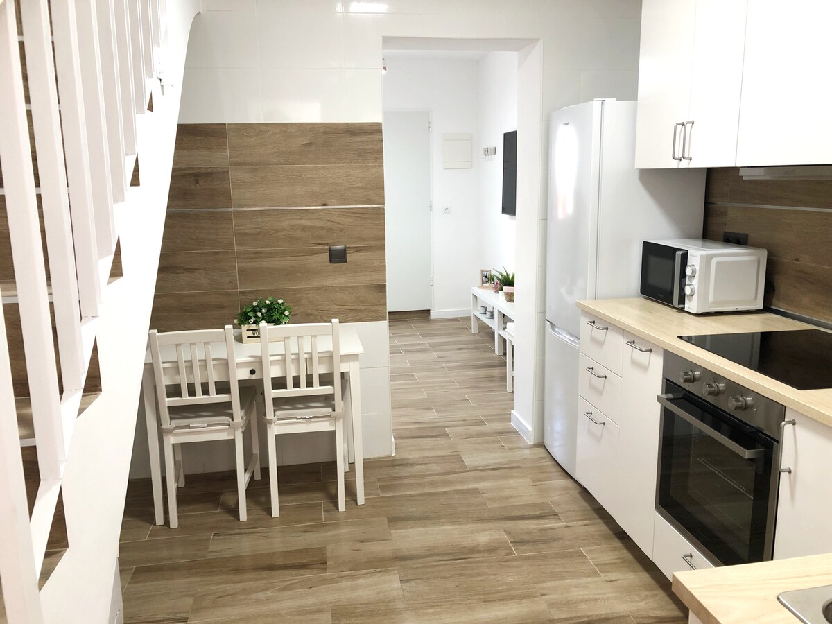 The kitchen area features contemporary white cabinetry paired with warm wooden accents. A small dining table with four chairs occupies one corner, while modern appliances, including a refrigerator and microwave, are visible. The floor tiles create a cohesive and spacious look throughout the area.
