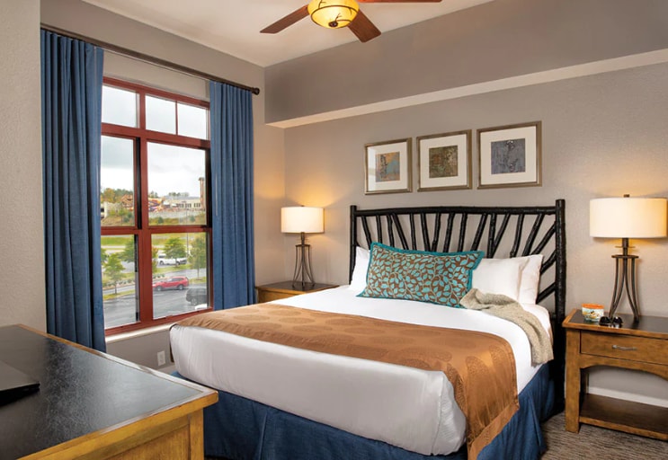 A well-lit bedroom features a spacious bed with a brown throw blanket and decorative cushions. Two bedside lamps provide gentle illumination. Light blue curtains frame a large window, enhancing the airy atmosphere. The wall displays three framed art pieces, adding a touch of character.