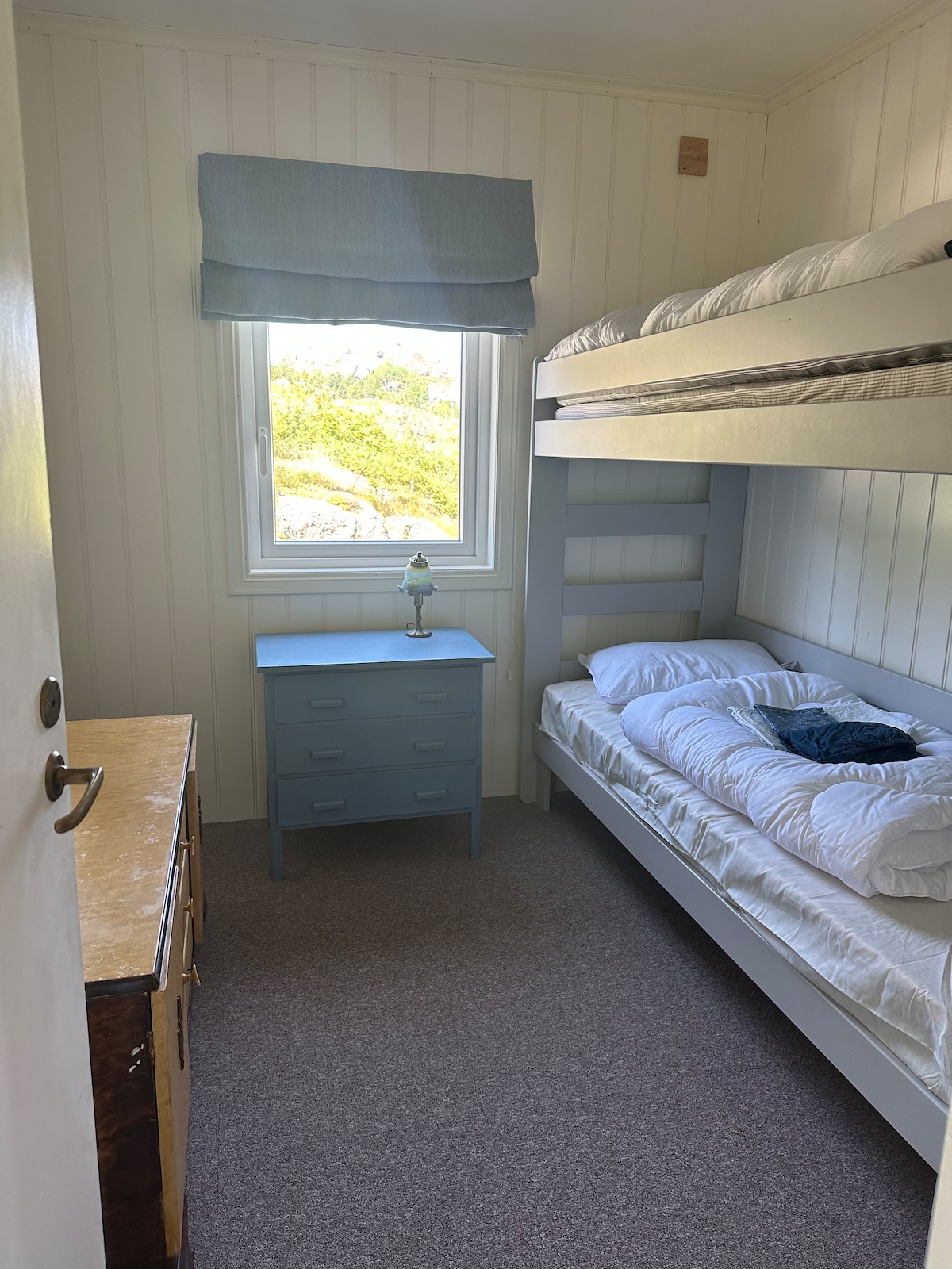 A well-lit bedroom features a bunk bed with a neatly arranged top and bottom mattress. A small blue dresser is positioned next to the bed. A table lamp sits on a bedside table near a window that offers views of greenery outside.