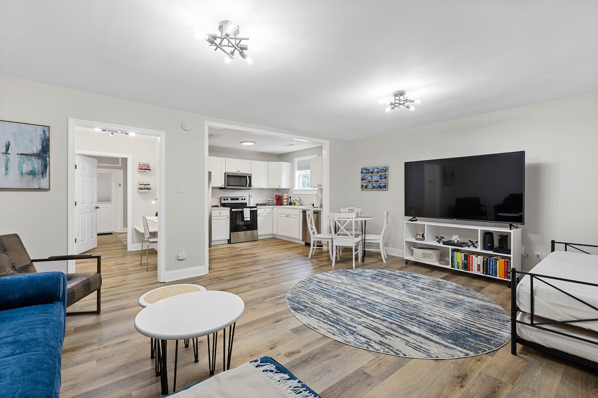 A spacious living area features a blue sofa and a white coffee table on a patterned rug. The room includes a large television mounted on a stand, with light wooden flooring flowing into the nearby kitchen. Bright overhead lights illuminate the area.