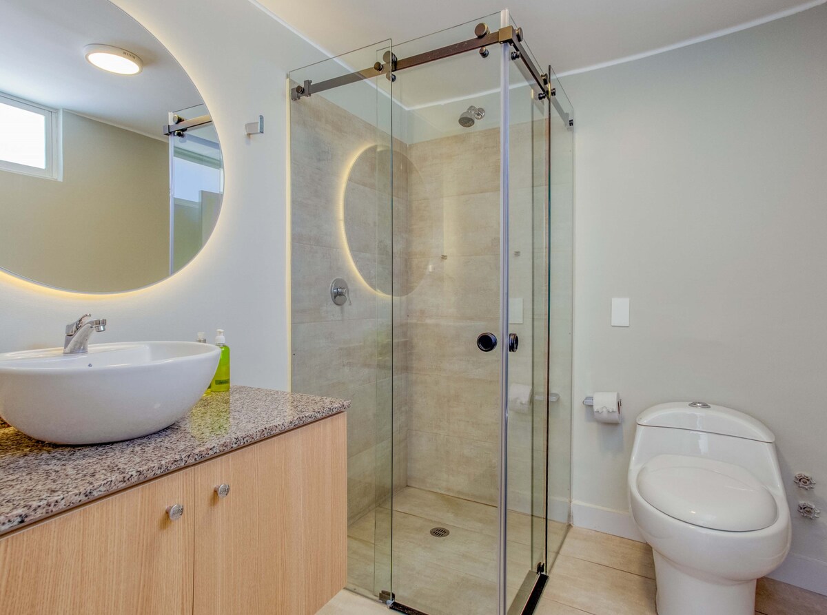A modern bathroom features a glass-enclosed shower with a sleek design. A round mirror is illuminated above a stone countertop, which holds a vessel sink. The clean lines of the space are complemented by neutral-colored walls and light flooring.