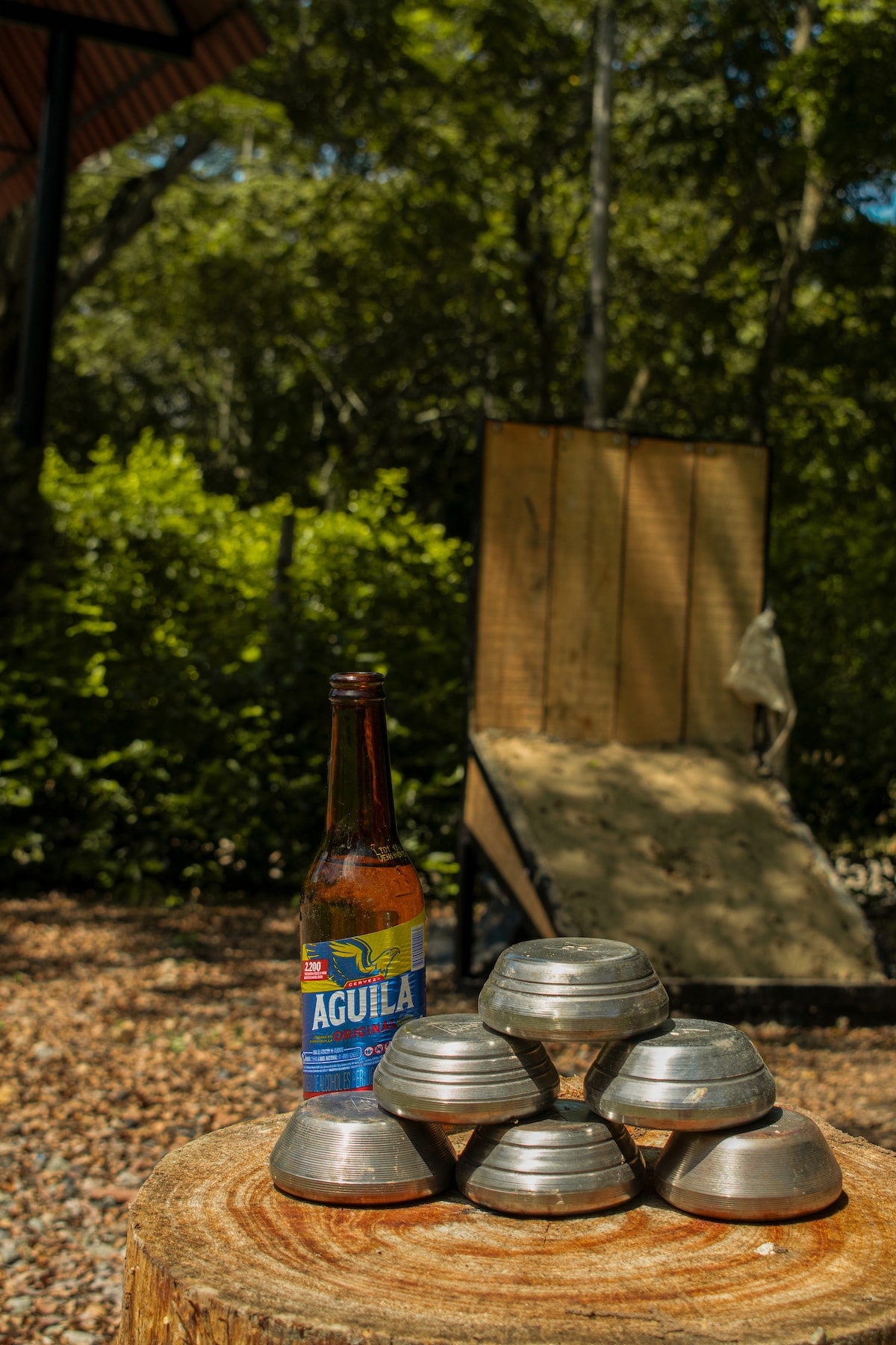 A collection of silver playing discs sits atop a wooden stump, while a bottle of Aguila beer is nearby. In the background, a wooden slope for games is visible, surrounded by lush greenery that provides shade and a sense of privacy.