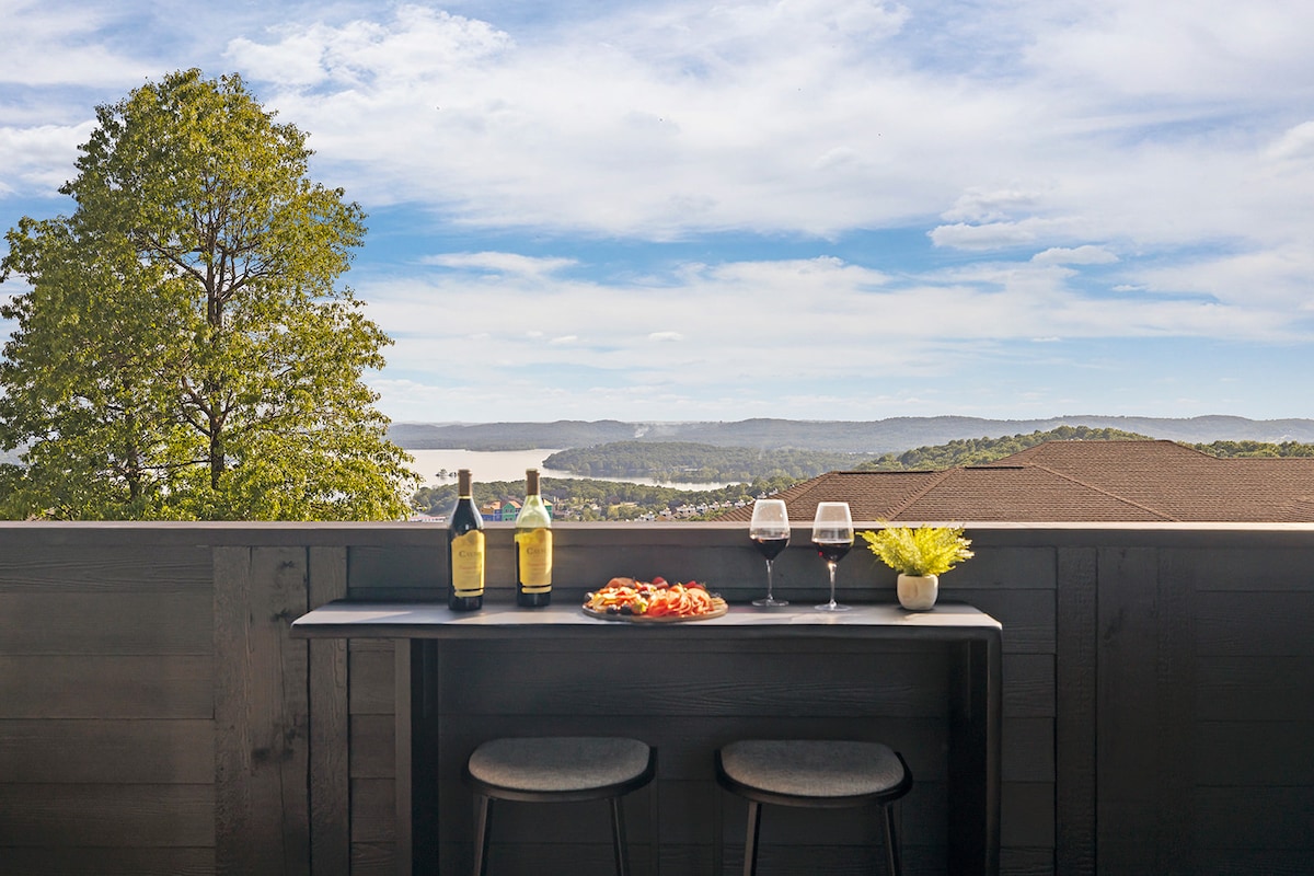A private balcony table is set for two, featuring wine bottles and glasses alongside a small plate of appetizers. Expansive views of Table Rock Lake and rolling hills are visible under a partly cloudy sky, providing a serene backdrop.