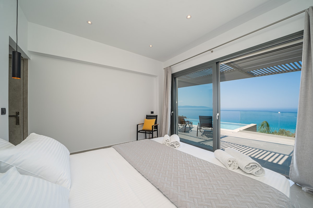A spacious bedroom features a large window offering views of the sea, allowing natural light to fill the room. The bed is dressed in white linens with a textured gray throw. A minimalist chair and table are positioned nearby, enhancing the room's simplicity.