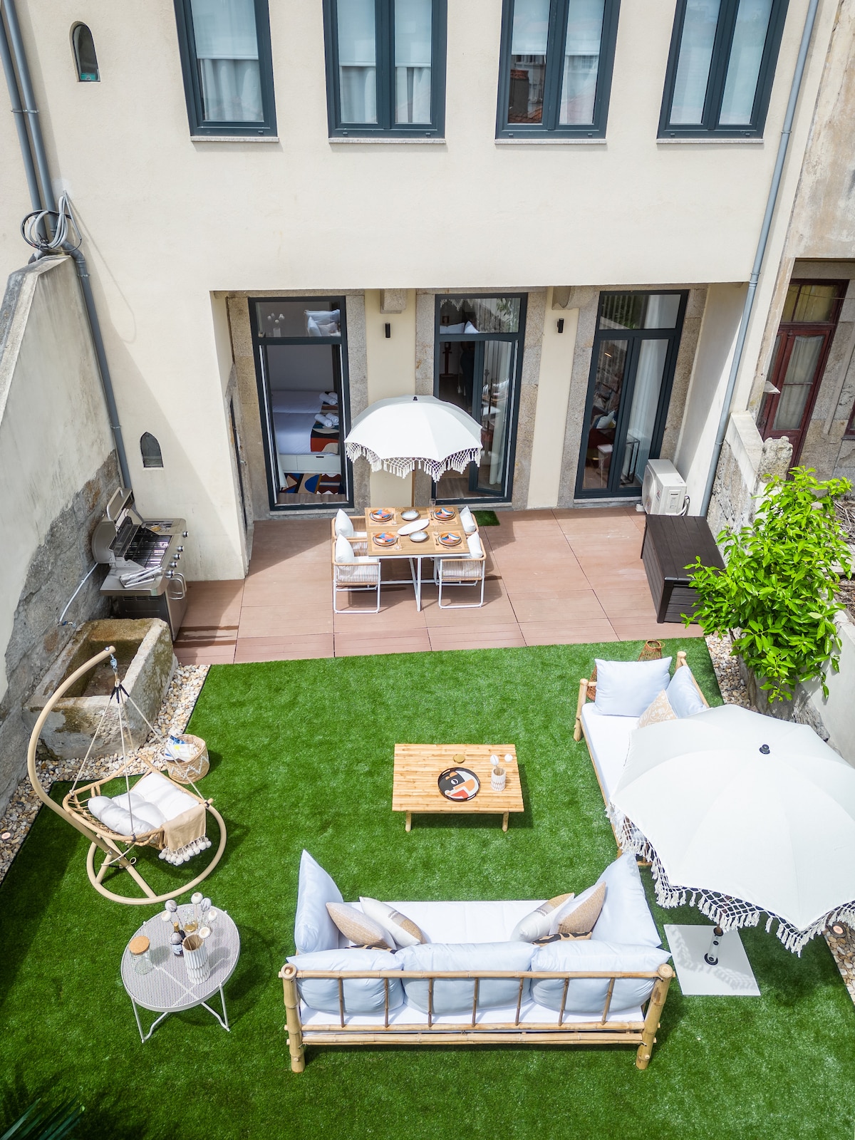 An outdoor garden is featured, showcasing a comfortable seating area with a light-colored sofa and decorative cushions. A dining table with chairs is beneath an umbrella, while a BBQ grill stands nearby. Lush green grass provides a vibrant backdrop, enhancing the space.