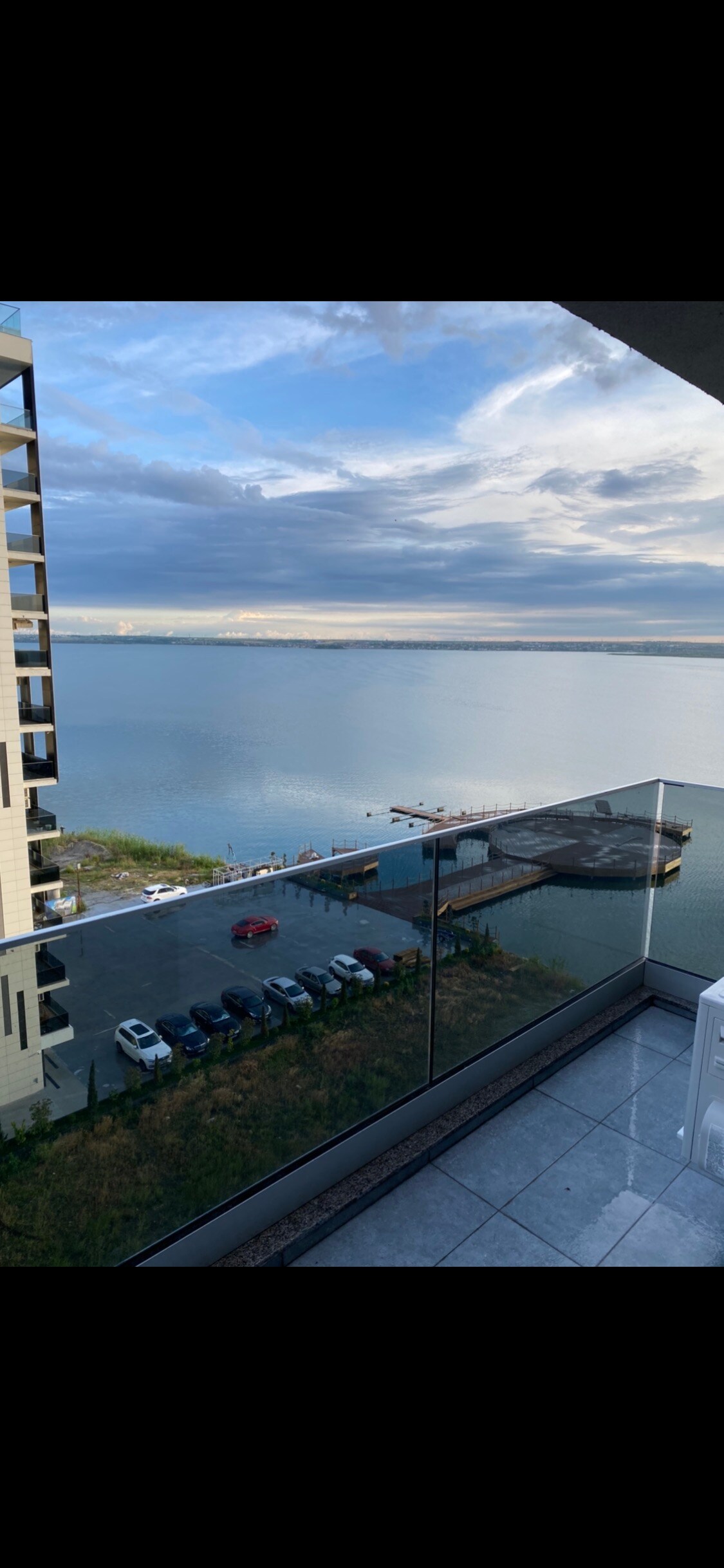 A panoramic view of the tranquil water is captured from the balcony, showcasing reflections of clouds and light on the surface. A parking area with several cars is visible below, framed by the sleek glass railing of the balcony.