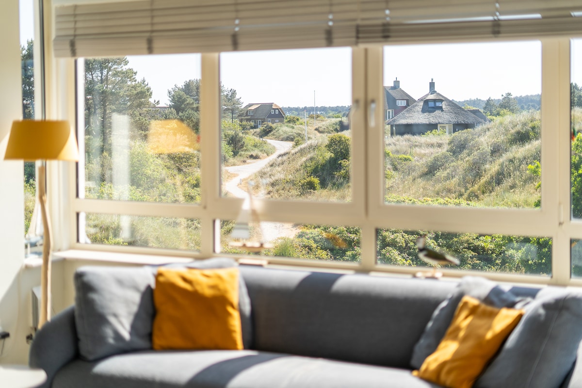 A spacious living area is viewed through large windows, revealing a serene landscape along a winding path. A soft grey sofa with yellow cushions enhances the comfortable ambiance, while the natural surroundings include clusters of greenery and distant houses.