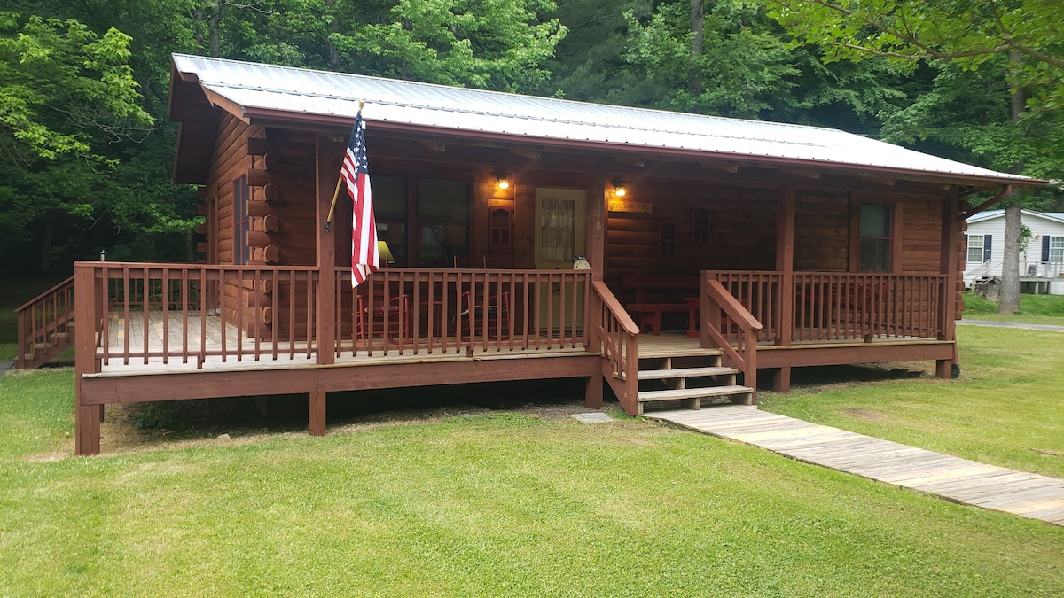 A cozy log home is surrounded by lush greenery, featuring a spacious deck with railings and steps leading up to the entrance. American flags are displayed, emphasizing the welcoming presence of this retreat in nature. The rustic exterior complements the serene environment.
