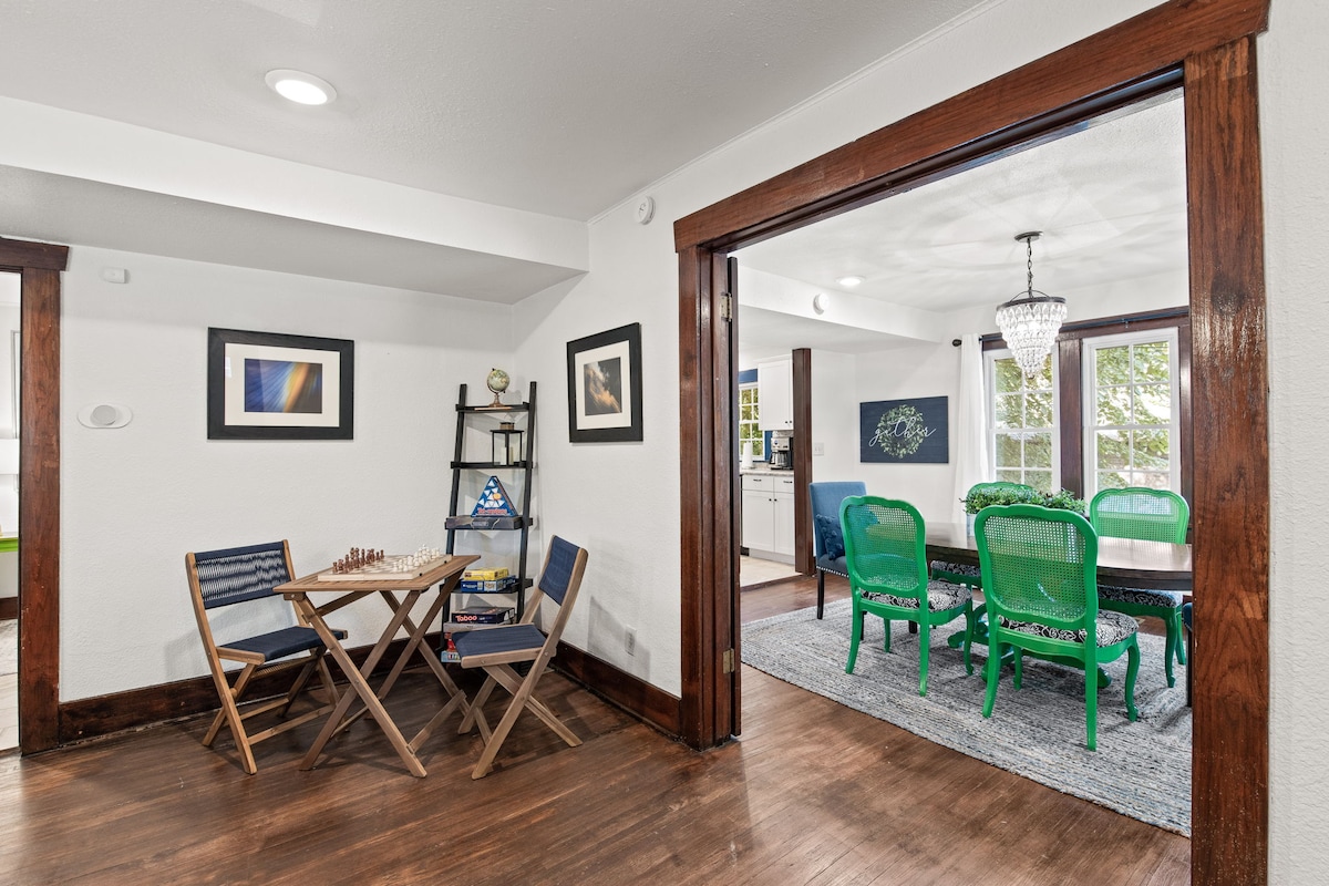 A cozy nook is presented with a small folding table and two matching chairs, set against a backdrop of white walls and dark wood accents. A chessboard is visible on the table, and a bookshelf stands nearby, adding to the inviting ambiance.