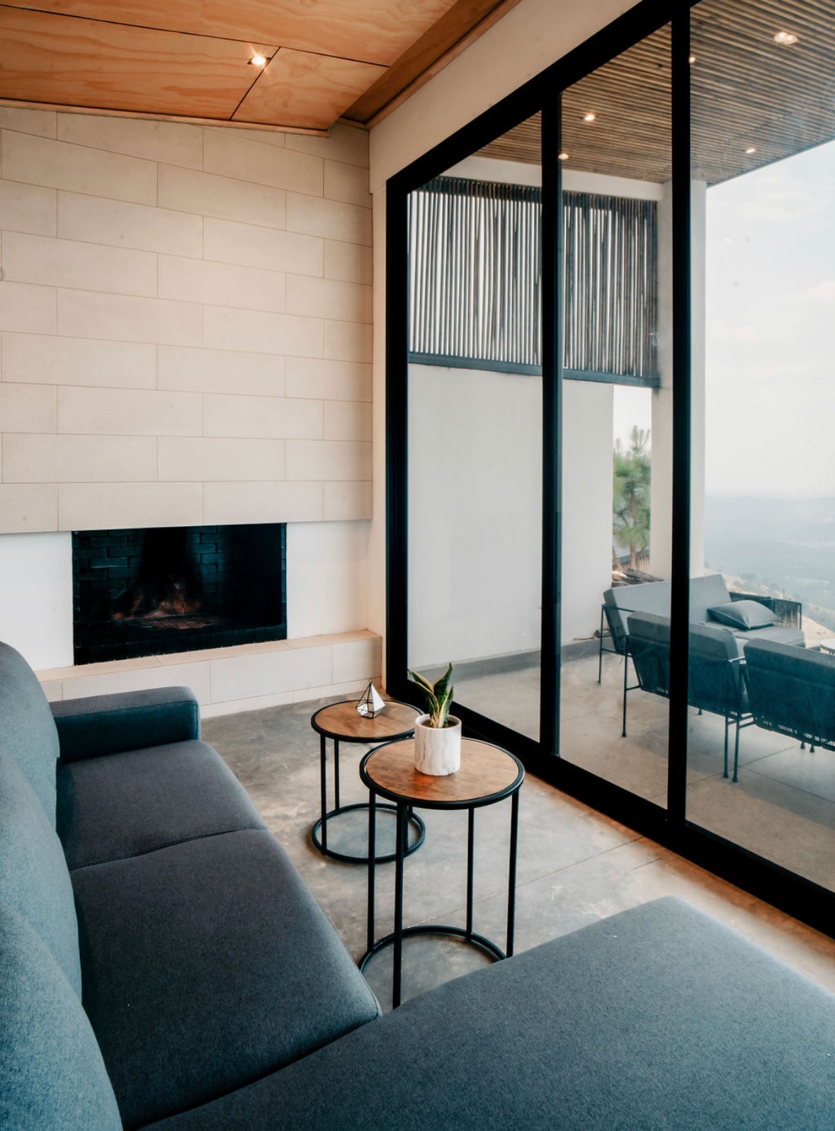 A modern seating area is featured with a comfortable gray sofa positioned beside two small tables. Large glass doors provide a view of the outdoors, while a minimalist fireplace enhances the space. Natural light fills the room, creating a serene environment.