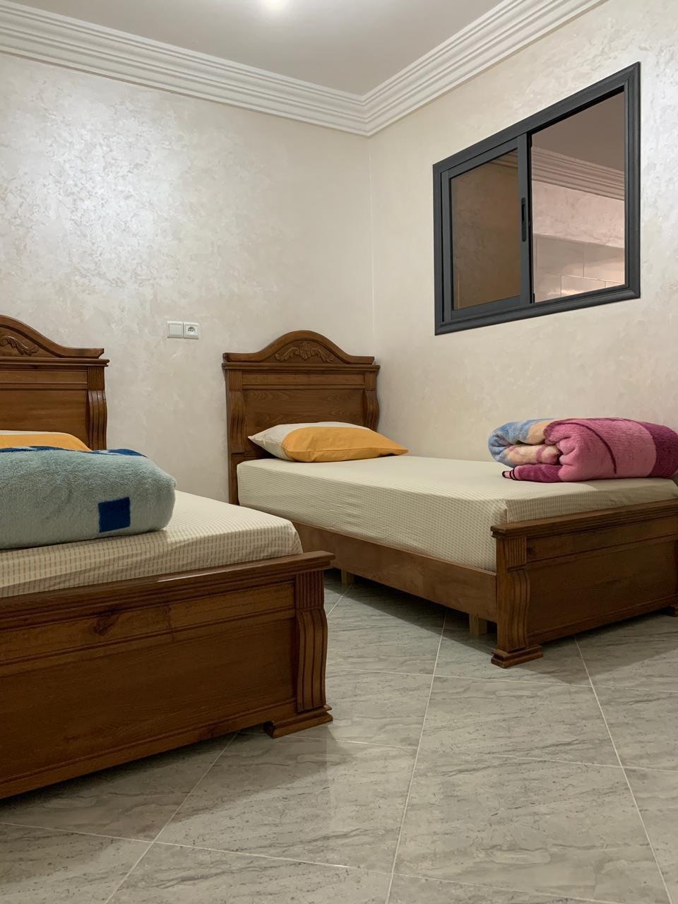 A room features two wooden beds, each dressed with light-colored bedding. A colorful rolled towel is placed at the foot of one bed. A large window allows natural light to enter, while the floor is covered with light-colored tiles.