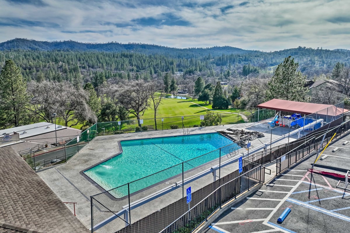 The image captures a large outdoor swimming pool surrounded by a fenced area. Lush green hills and trees are visible in the background, while an 18-hole golf course extends into the distance. A shaded area with playground features can be seen nearby.