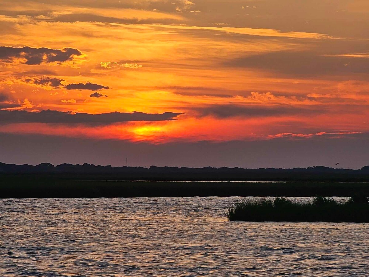 A vibrant sunset casts warm hues over a calm body of water, with silhouettes of distant trees visible along the horizon. The sky is filled with clouds that reflect orange and gold tones, creating a serene atmosphere.