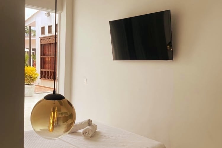 A light and airy room is showcased with a spacious bed covered in white linens. A modern television is mounted on the wall opposite. A stylish pendant light hangs from the ceiling, providing a warm glow, while natural light filters in from an open door leading to an outdoor area.