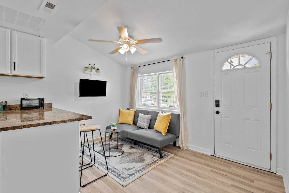 The inviting living area features a comfortable gray sofa adorned with yellow cushions. A small round dining table with two chairs is positioned beside the kitchenette, while a wall-mounted TV offers entertainment options. Natural light enters through the window next to the front door.