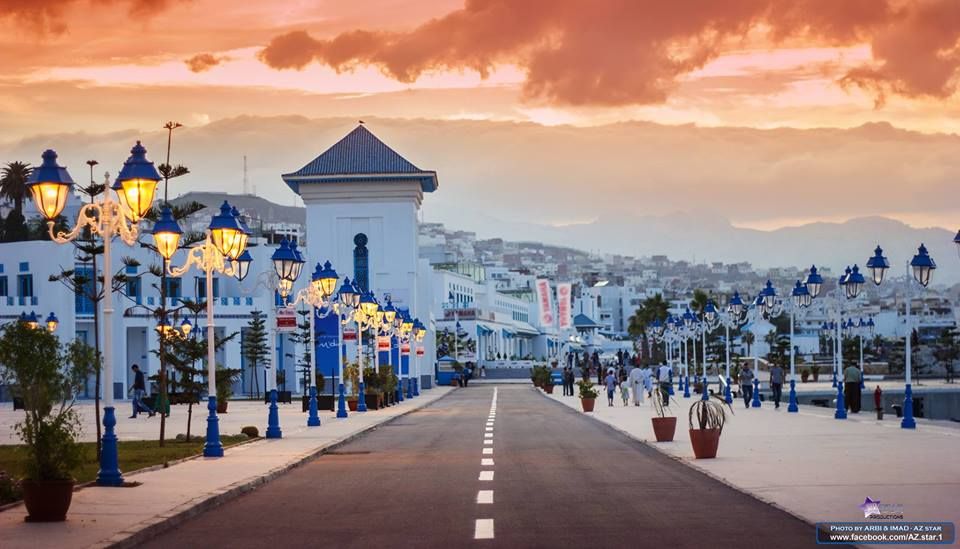 A waterfront promenade is depicted under a vibrant sky at dusk. Decorative streetlamps line the path, which is flanked by buildings featuring a mix of white and blue architecture. People can be seen strolling along the walkway, contributing to a lively atmosphere.
